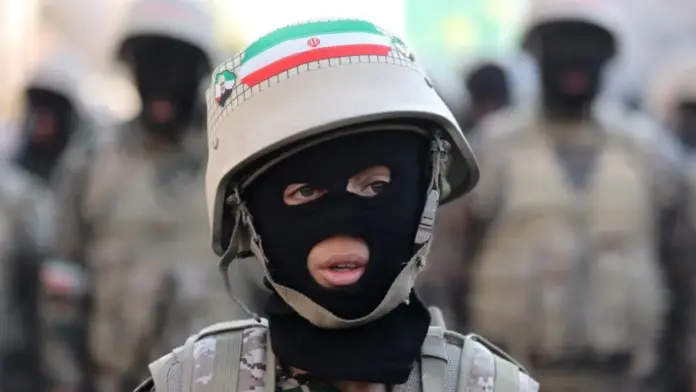 Majid Asgaripour/WANA/Reuters A young person wearing a balaclava, fatigues and a helmet sporting an Iranian flag looks off camera, with blurred military figures in the background.
