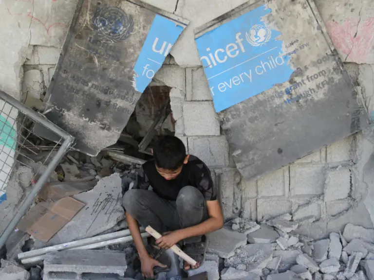 A boy sits under a broken UNICEF sign as Palestinians inspect the site of an Israeli strike on a school sheltering displaced people [File: Mahmoud Issa/Reuters]