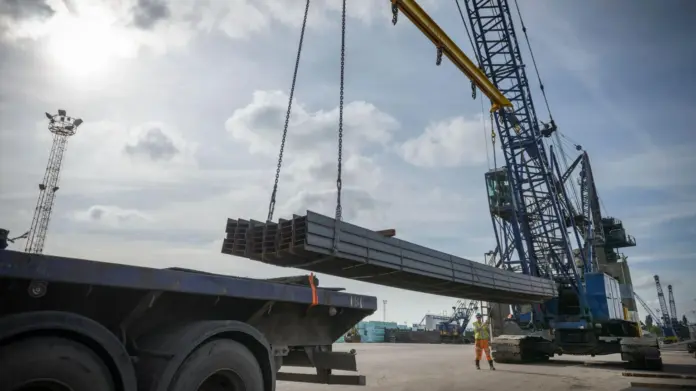 BBC Large steel rods are lifted with a crane. It is a sunny day with blue skies.