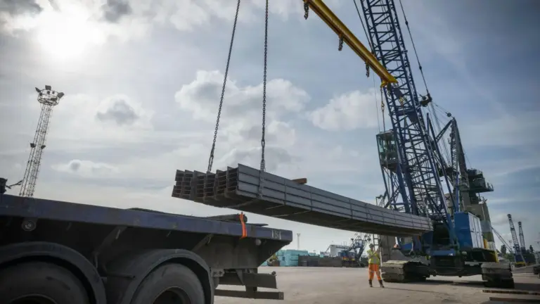 BBC Large steel rods are lifted with a crane. It is a sunny day with blue skies.